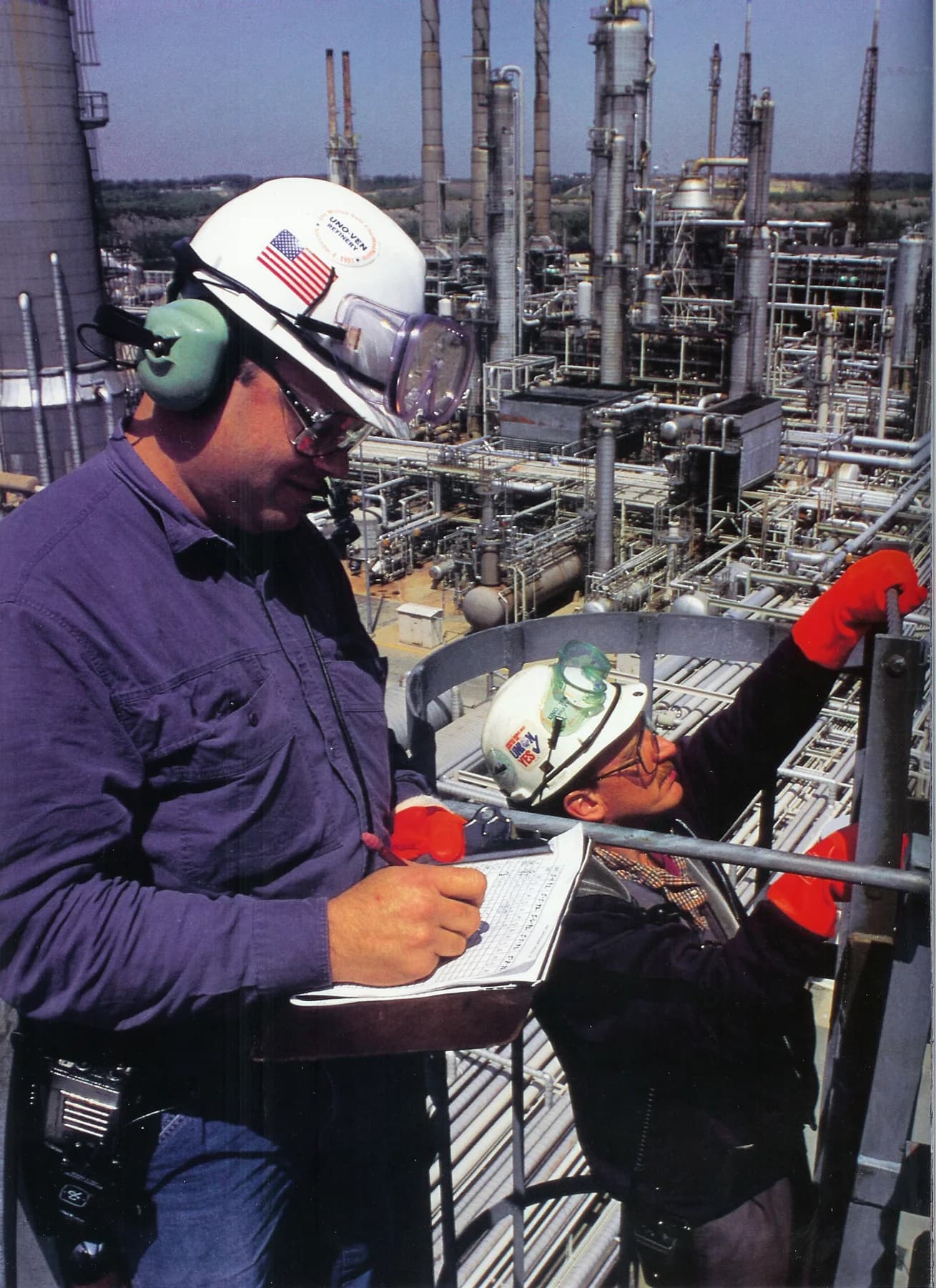 Two workers in hard hats reviewing documentation on a refinery tower platform, archival photograph